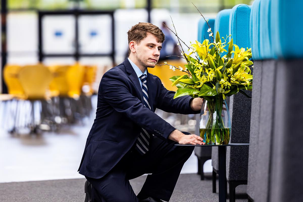 Male front of house employee looking after a vase of yellow flowers on a table in reception area